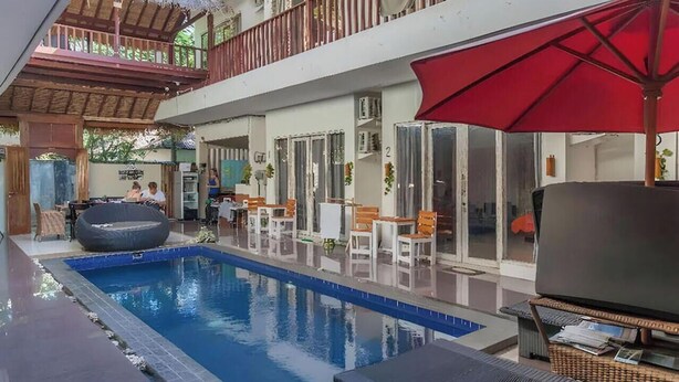 An indoor swimming pool with lounge chairs and umbrellas, a balcony with a thatched roof, and a second-floor balcony with railings. - CJs House Gili Trawangan (Gili Trawangan)