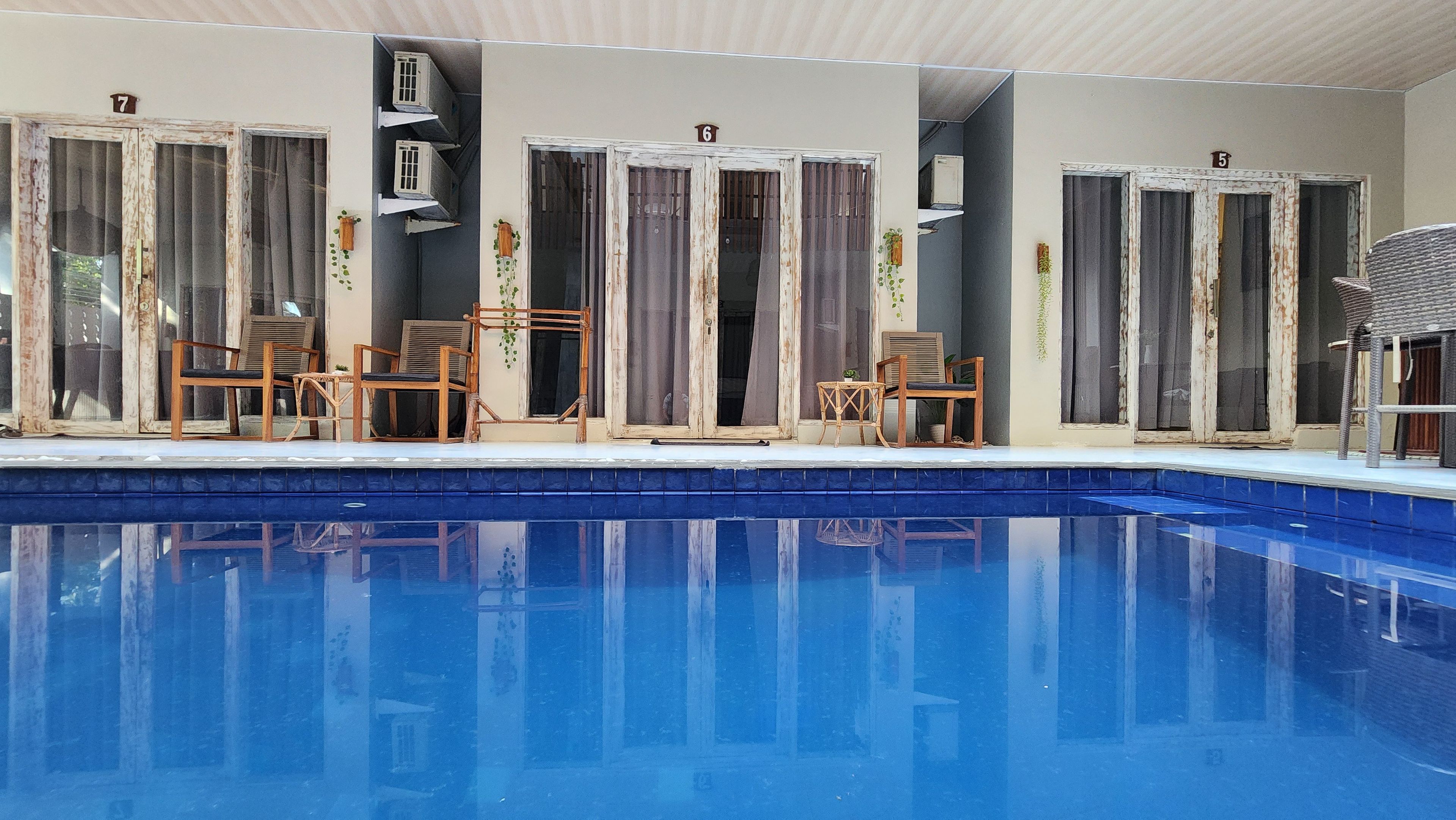 A swimming pool with a tiled edge, surrounded by wooden chairs and tables, and a series of doors with numbers on them.