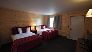 A room with two beds, wooden walls, a window with curtains, a door, and a desk with a lamp and remote controls.