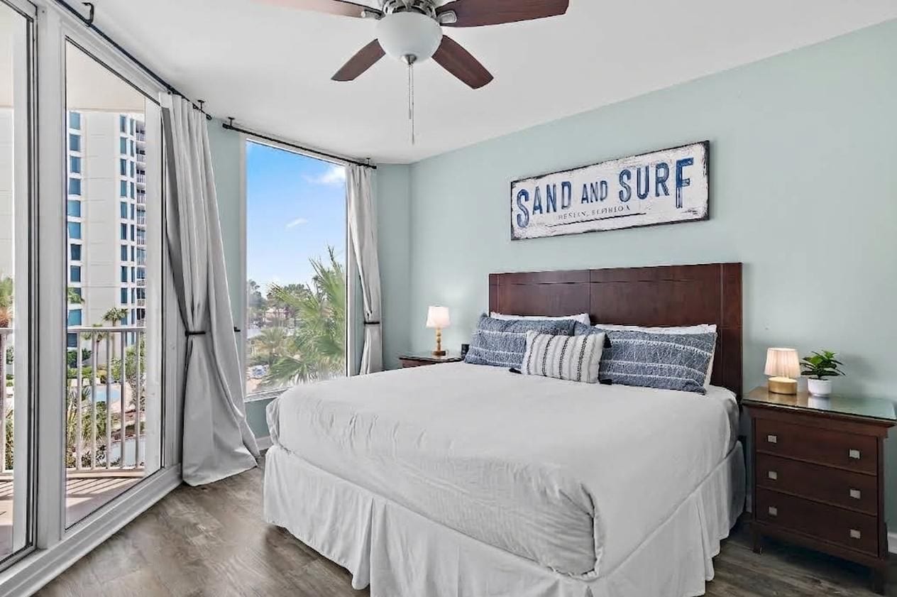 Een slaapkamer met een groot bed, een plafondventilator, uitzicht op palmbomen en een bord met de tekst 'SAND AND SURF'.