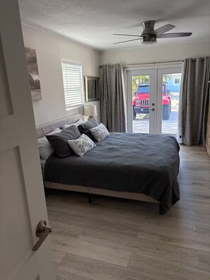 Ein Schlafzimmer mit Bett, Deckenventilator, Fenster mit Jalousien und einer Tür, die nach draußen führt.
