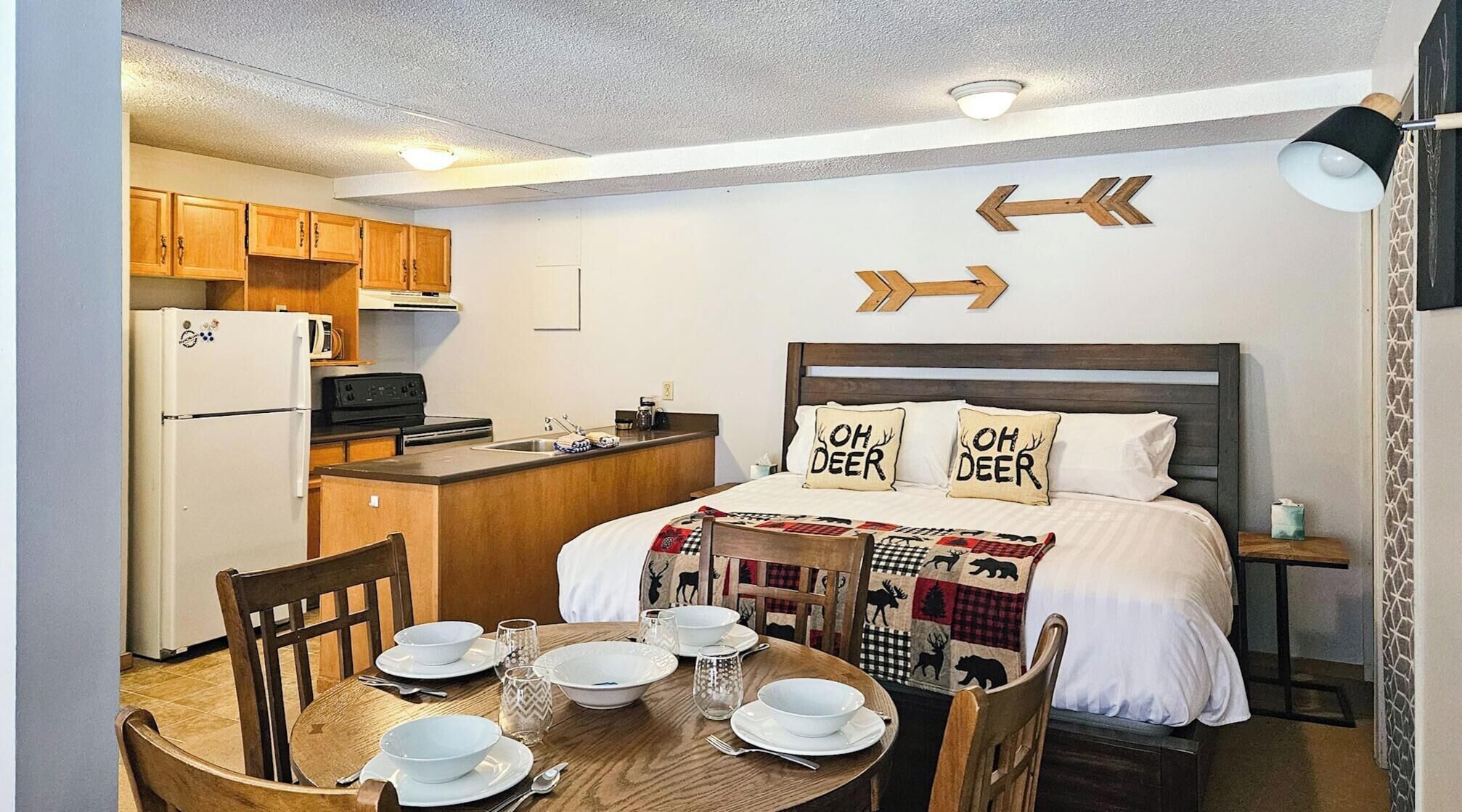 A compact hotel room with a kitchenette, a bed with deer-themed pillows, a dining table set for two, and directional arrows on the wall.