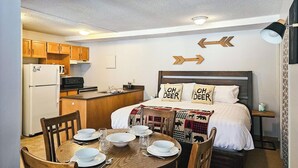A compact hotel room with a kitchenette, a bed with deer-themed pillows, a dining table set for two, and directional arrows on the wall. - King Studio / Hot Tubs, Pools & Ski Access (Panorama)