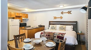A compact hotel room with a kitchenette, a bed with deer-themed pillows, a dining table set for two, and directional arrows on the wall.