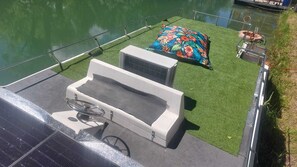 Un bateau doté d’un petit espace de pont comprenant une baignoire blanche, une couverture à motifs floraux colorés et un petit coin salon avec une chaise et un gilet de sauvetage.