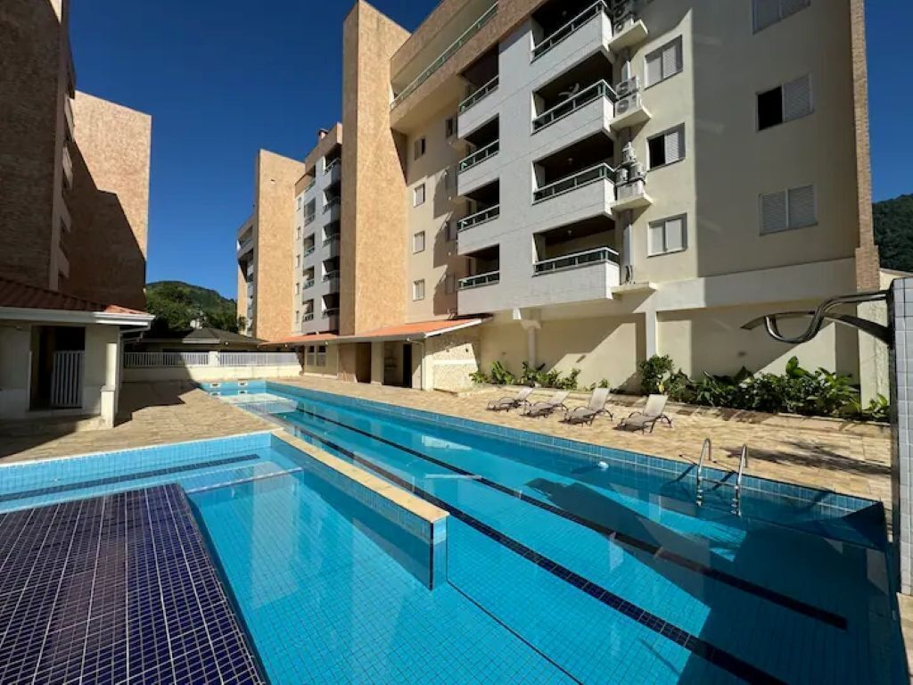 A swimming pool with adjacent sun loungers, a building with balconies, and a clear blue sky.