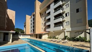 A swimming pool with adjacent sun loungers, a building with balconies, and a clear blue sky. - Casa Melo 8 - Ubatuba (Ubatuba)