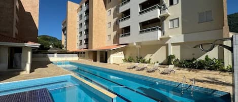 A swimming pool with adjacent sun loungers, a building with balconies, and a clear blue sky.