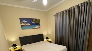 A bedroom with a bed, a ceiling fan, a nightstand with a lamp, a painting of a beach scene, and a window with curtains. - Casa Melo 8 - Ubatuba (Ubatuba)