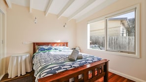 Une chambre avec un lit en bois, une table de chevet, une fenêtre donnant sur une clôture en bois et un plafond avec des poutres apparentes.
