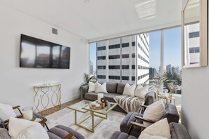 A modern living room with a large flat-screen TV, a sofa set, a coffee table, and a view of a cityscape through large windows.