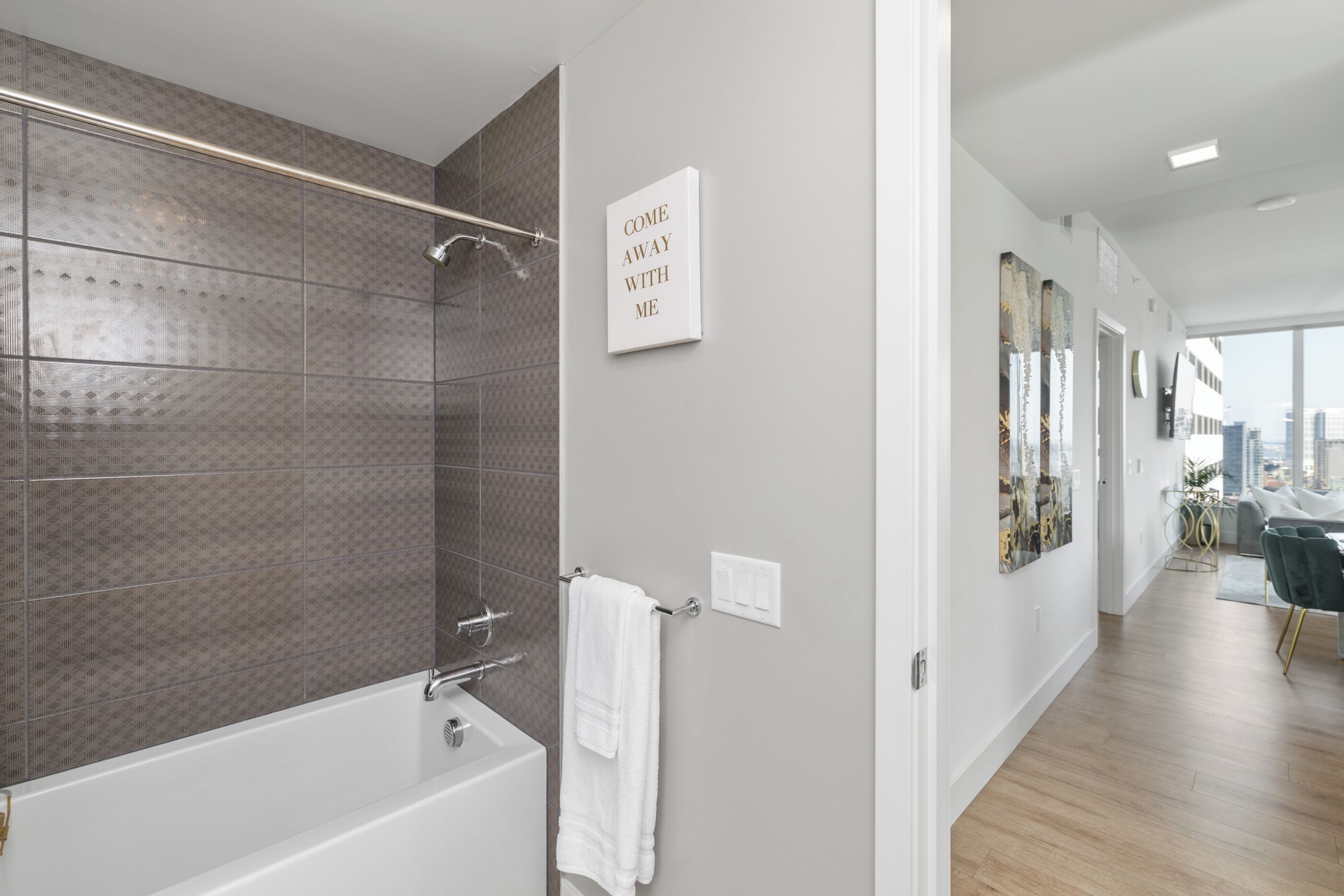 A modern bathroom with a bathtub, a shower, and a towel rack. There is a sign on the wall that reads "COME AWAY WITH ME". The bathroom has a tiled wall and a wooden floor.