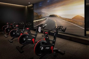 Een sportschool met hometrainers, een groot scherm dat een landschappelijk wegbeeld toont, en een donker interieur.