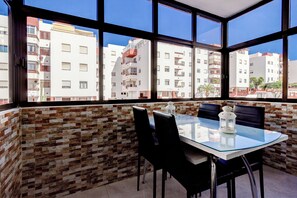 A dining area with a glass table and chairs, a white lantern, and a view of apartment buildings through large windows. - Ocean Breeze San Eugenio (Costa Adeje)