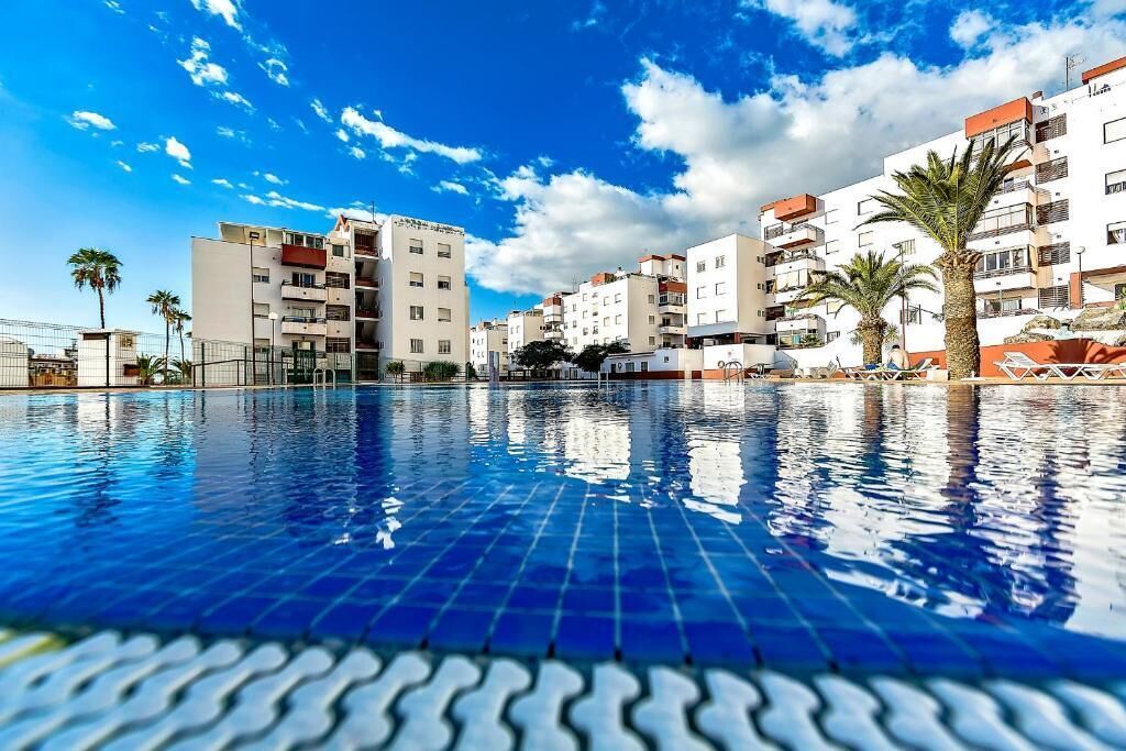 A swimming pool with tiled flooring, surrounded by multi-story buildings with balconies, palm trees, and a clear sky.