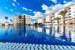 A swimming pool with tiled flooring, surrounded by multi-story buildings with balconies, palm trees, and a clear sky. - Ocean Breeze San Eugenio (Costa Adeje)