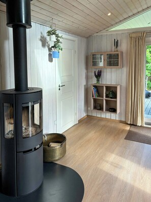 A modern interior with a wood-burning stove, a door, a shelf with decorative items, and a view through a window. - Covered terrace and beach at Fjellerup (Glesborg)
