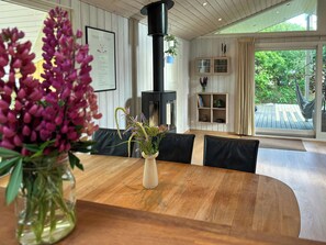 A dining area with a wooden table, black leather chairs, a vase with flowers, a black stove, and a bookshelf. - Covered terrace and beach at Fjellerup (Glesborg)