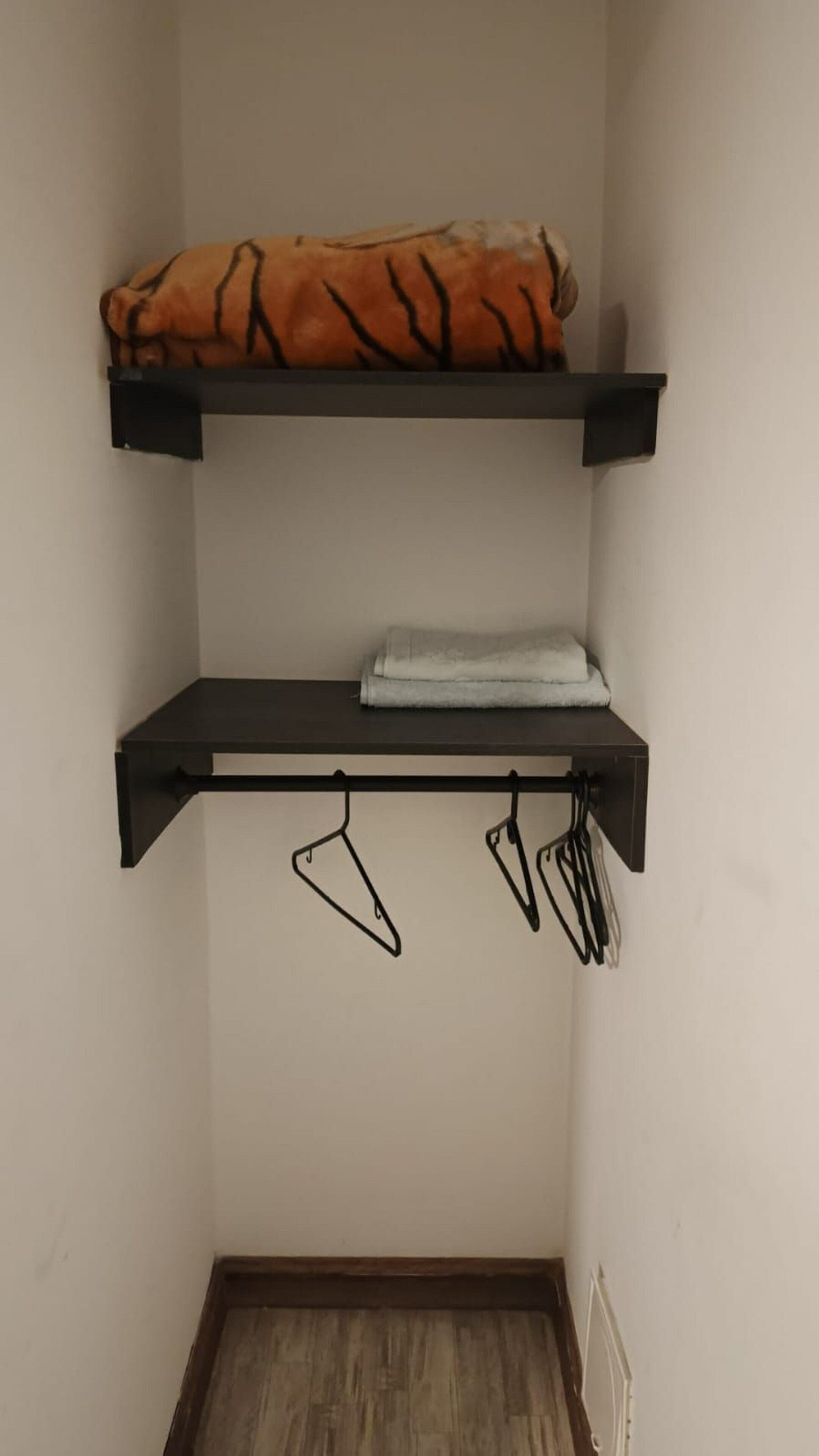 A room with two black shelves, one holding a folded orange and black blanket and the other with a white towel. Below, a black metal rack holds several black hangers.