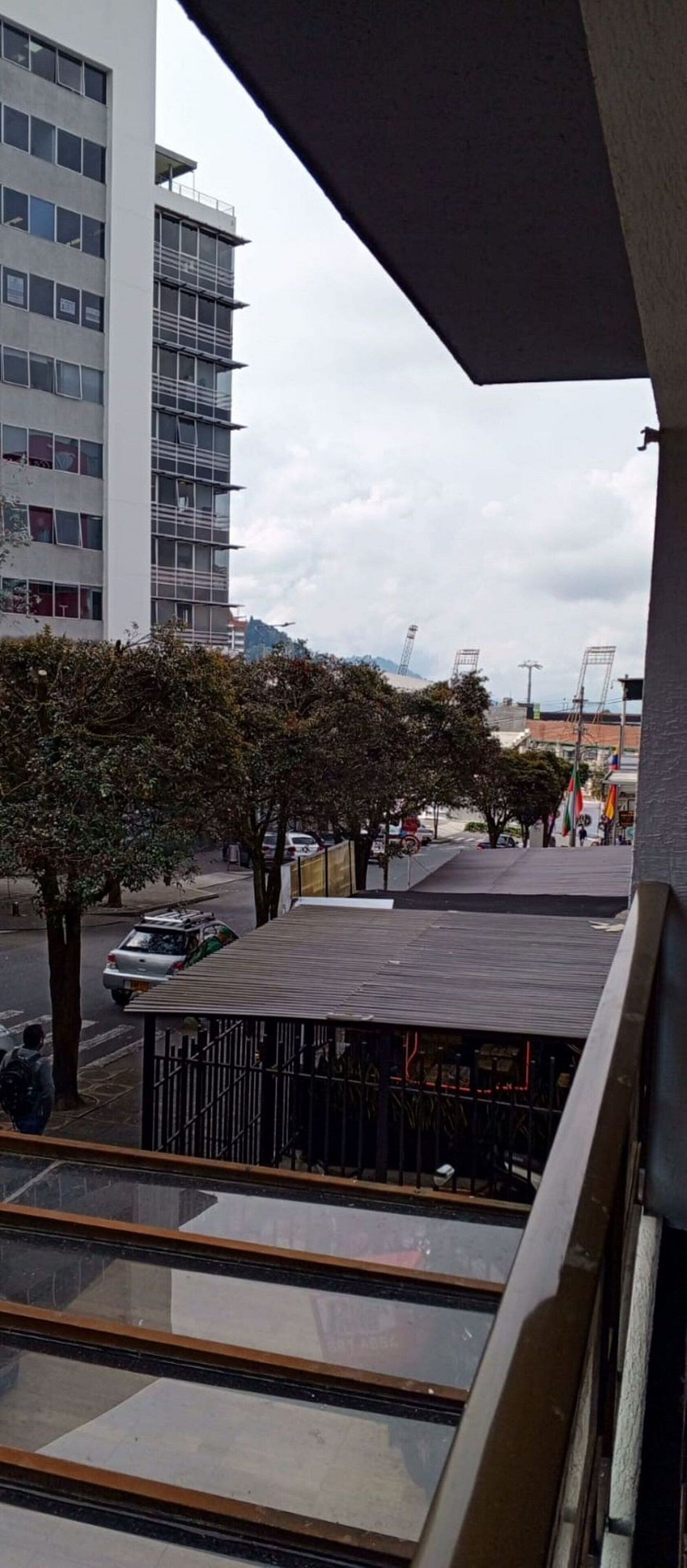 Vista de uma varanda com vista para uma rua com árvores, um carro estacionado e um prédio com andaimes.