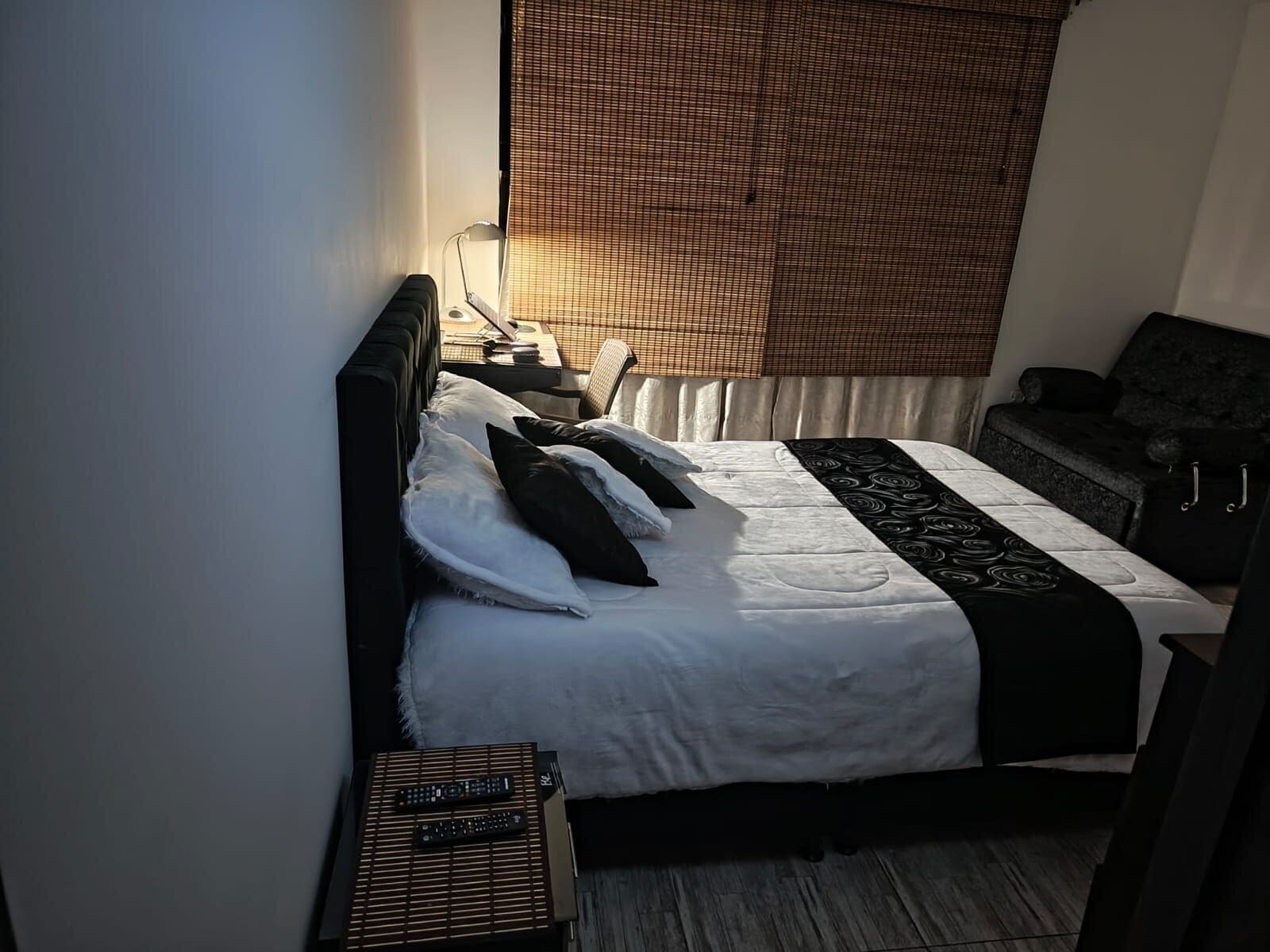 Quarto de hotel com cama, mesa de cabeceira, cadeira e janela com persianas.