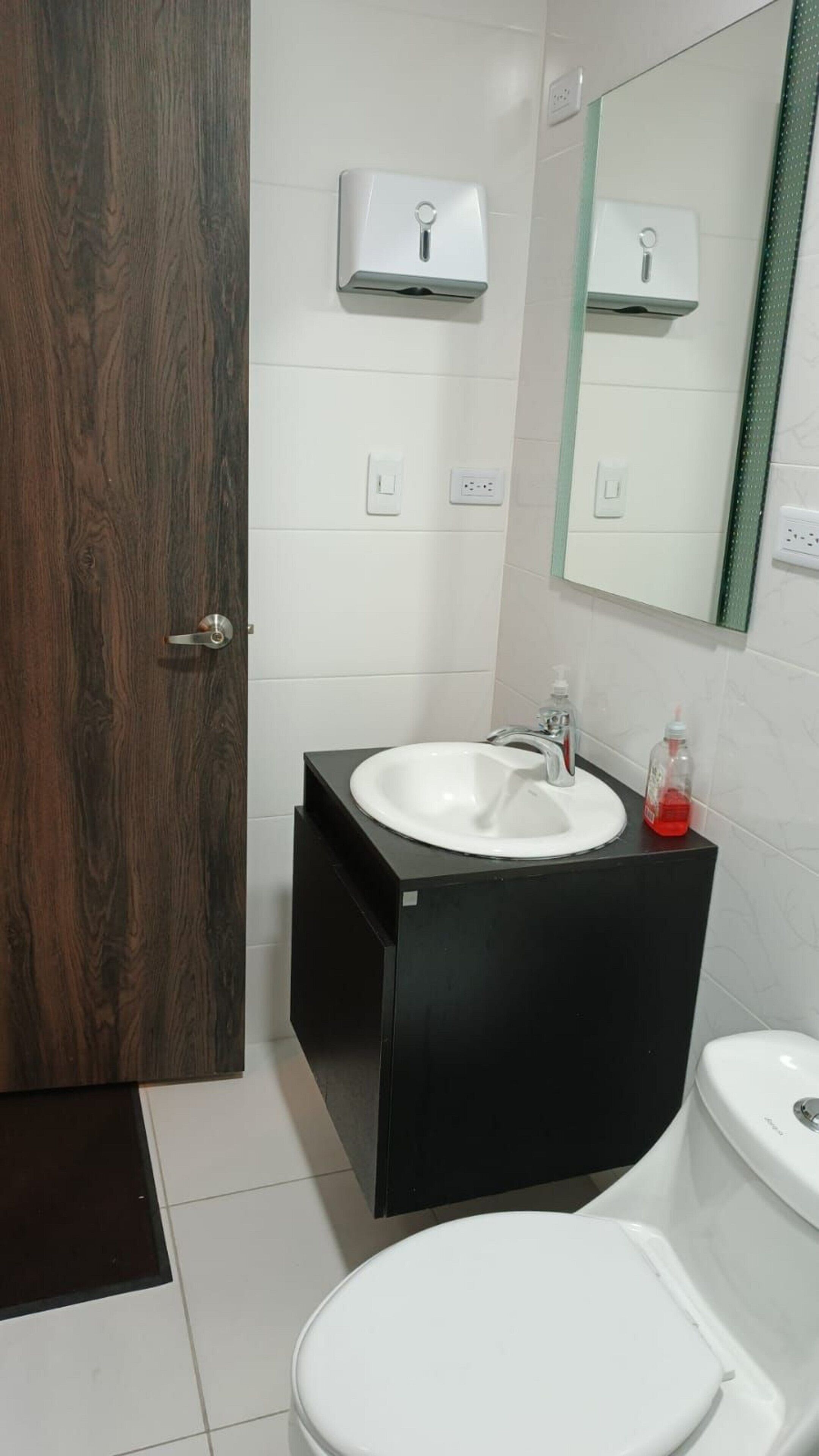 A bathroom with a wooden door, a white sink with a black countertop, a mirror above the sink, and a white toilet.