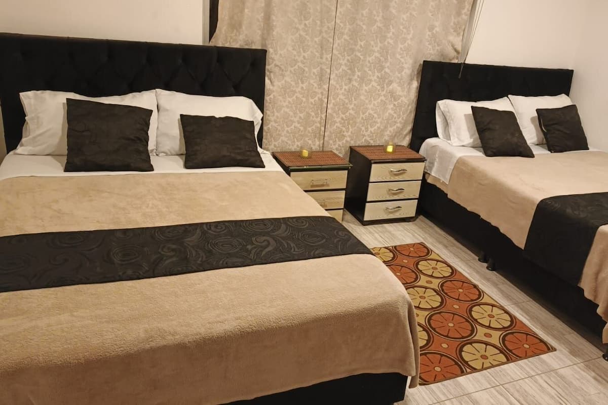 Ein Hotelzimmer mit zwei Betten, einem kleinen Nachttisch mit Lampe, einem gemusterten Teppich und einem Fenster mit Vorhängen.