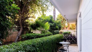 A patio with a table and chairs, surrounded by lush greenery and a tree. - Luxury Glyfada flat steps from sea & main market (Glifada)