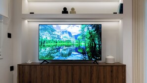 A flat-screen TV mounted on a wooden cabinet, with a magazine on a glass table in front. - Luxury Glyfada flat steps from sea & main market (Glifada)