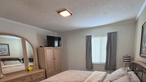 Ein Schlafzimmer mit einem Bett, einer Kommode, einem Fernseher auf einem Schrank, einem Fenster mit Vorhängen und einer Deckenleuchte.