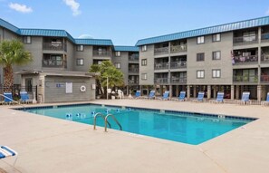 Una piscina con sillas de descanso, un pequeño edificio con un letrero y un edificio residencial de varios pisos con balcones.