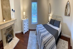 A bedroom with a fireplace, a bed with a striped bedspread, a nightstand with a lamp, and a window with blinds.