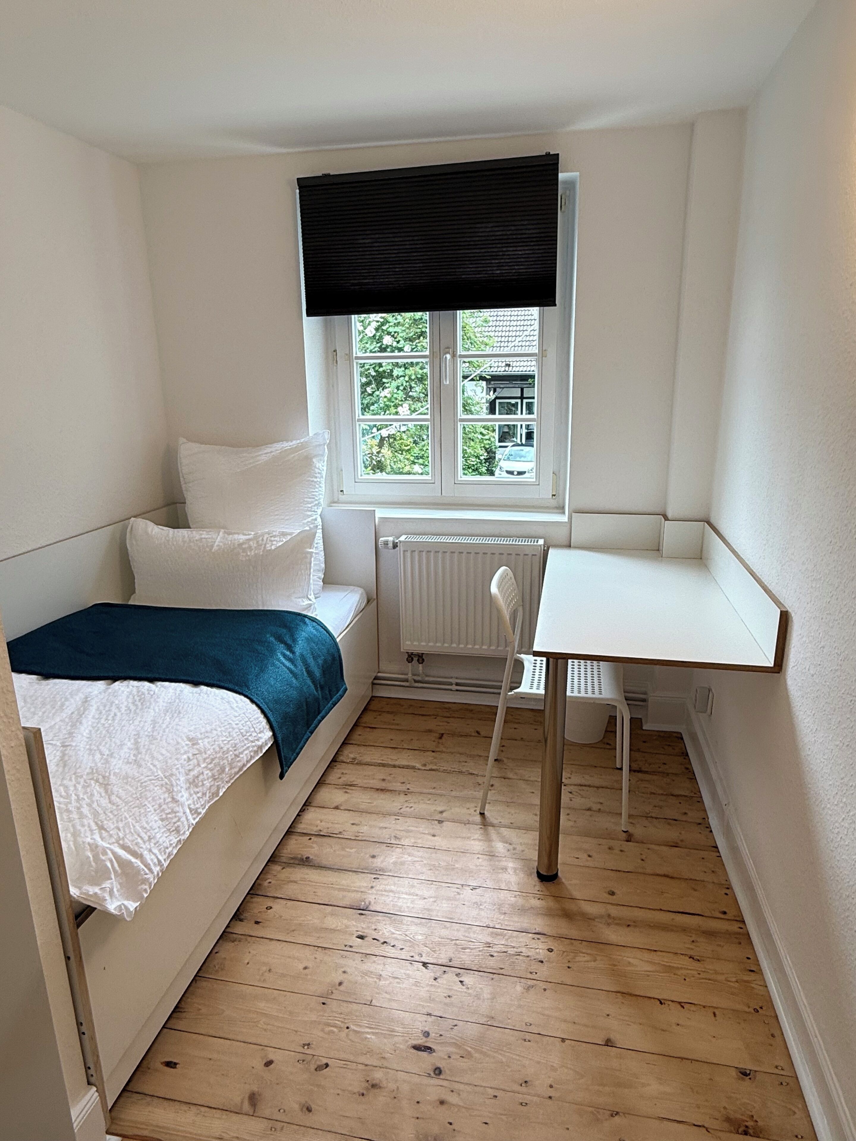A small, single-room apartment with a bed, a desk, and a window.