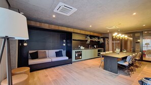 A modern kitchen with a central island, a dining area with a large table and chairs, a sofa with cushions, and a ceiling vent. - Harmonia da Vila (São Paulo)