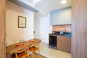 A compact kitchen with wooden cabinets, a black refrigerator, and a dining area with a wooden table and orange chairs. - Harmonia da Vila (São Paulo)