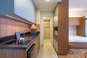 A compact kitchen with a sink, stove, and microwave, leading to a small bedroom with a single bed and a chair. - Harmonia da Vila (São Paulo)