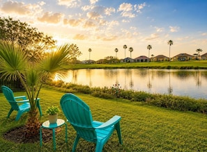 A serene lakeside setting with a row of houses, a palm tree, and two turquoise chairs.