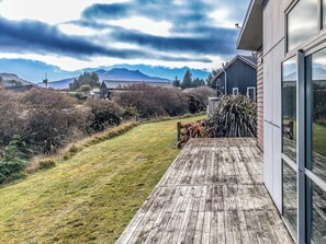 A wooden deck leading to a modern building with large windows, a grassy yard, and mountains in the background. - Mountainview Glimpse - Ohakune Holiday Home (Ohakune)