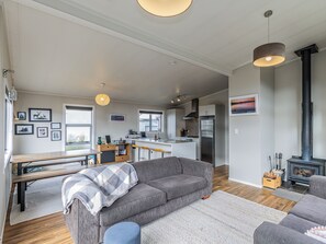 A modern living space with a grey sofa, a wooden dining table, a kitchen area, and a wood-burning stove. - Mountainview Glimpse - Ohakune Holiday Home (Ohakune)