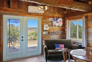A rustic cabin interior with a wooden ceiling, a couch with an American flag pillow, a dining table, and a view of the outdoors through large windows. - Charming rustic 1 bedroom cabin (Prescott)