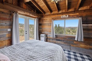 A wooden cabin room with a bed, a window with curtains, a sign reading 'Roses,' and a view of the outdoors. - Charming rustic 1 bedroom cabin (Prescott)