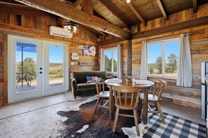 A rustic cabin interior with wooden walls, a dining area with a round table and chairs, a sofa with an American flag pillow, and large windows with curtains. - Charming rustic 1 bedroom cabin (Prescott)