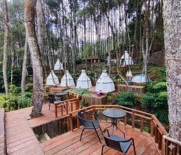 A wooden deck with outdoor seating, surrounded by trees and multiple conical tents. - Cozzy Saung Singkur Riverside (bandung)