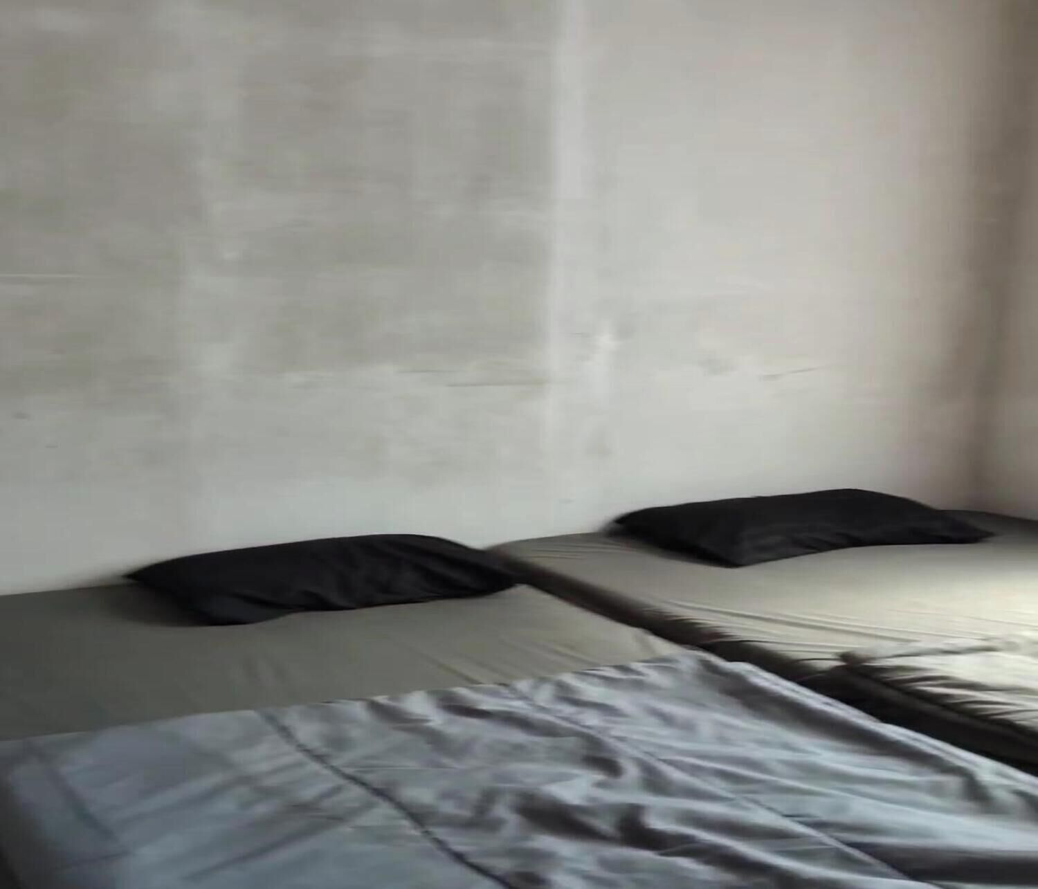 A room with two beds, each with a black pillow. The walls are plain and light-colored.