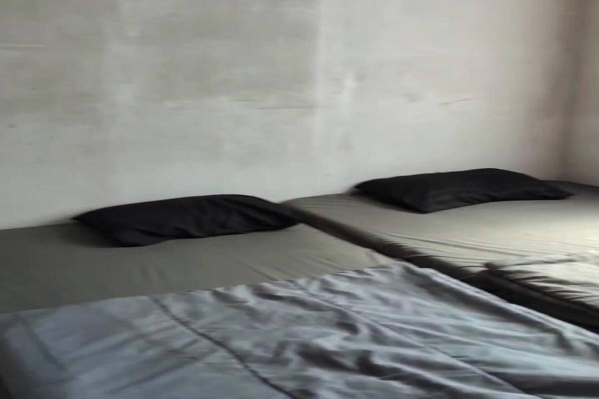 A room with two beds, each with a black pillow. The walls are plain and light-colored.