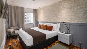 Una habitación de hotel moderna con cama, mesita de noche, lámpara y vistas a la ciudad por la ventana.