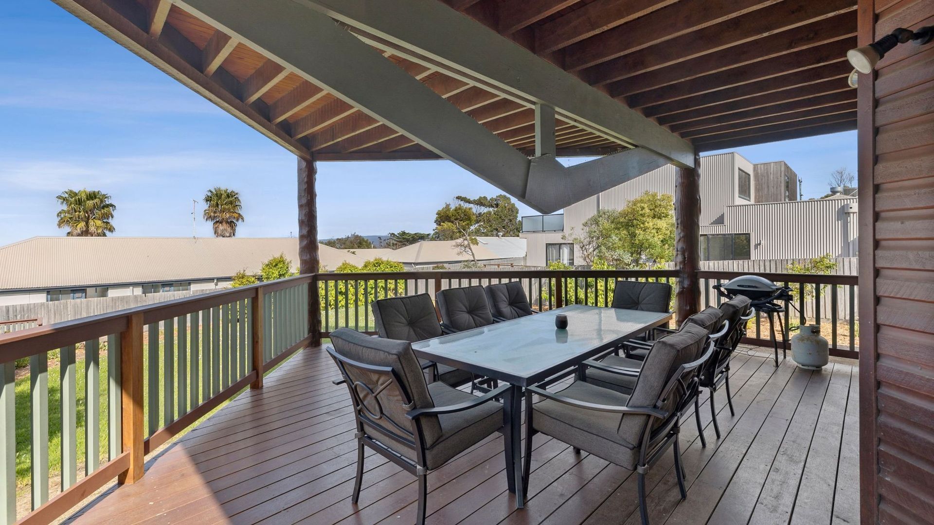 A covered outdoor deck with a dining table and chairs, a grill, and a view of the neighborhood.