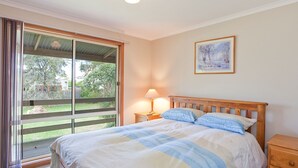 A bedroom with a bed, a nightstand, a lamp, a window with curtains, and a painting on the wall. - The Sundowner Cottage - Central Torquay (Torquay)