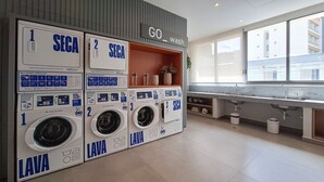 A laundry area with multiple washing machines, a sink, and a window with a view of buildings. - Nex One Paraiso (São Paulo)