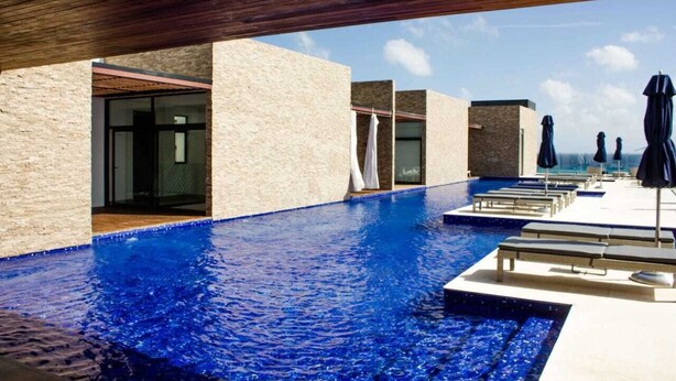 A modern pool area with clear blue water, sun loungers, and umbrellas, surrounded by stone walls and wooden beams. - Beautiful One-Bedroom, Two-Full-Bathroom Condo Just Steps from the Beach! (Playa del Carmen)